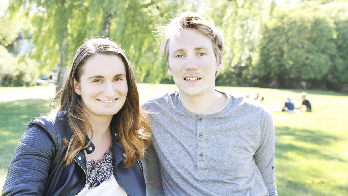 Två studenter sitter i gröngräset utanför universitetet. De ler mot kameran.