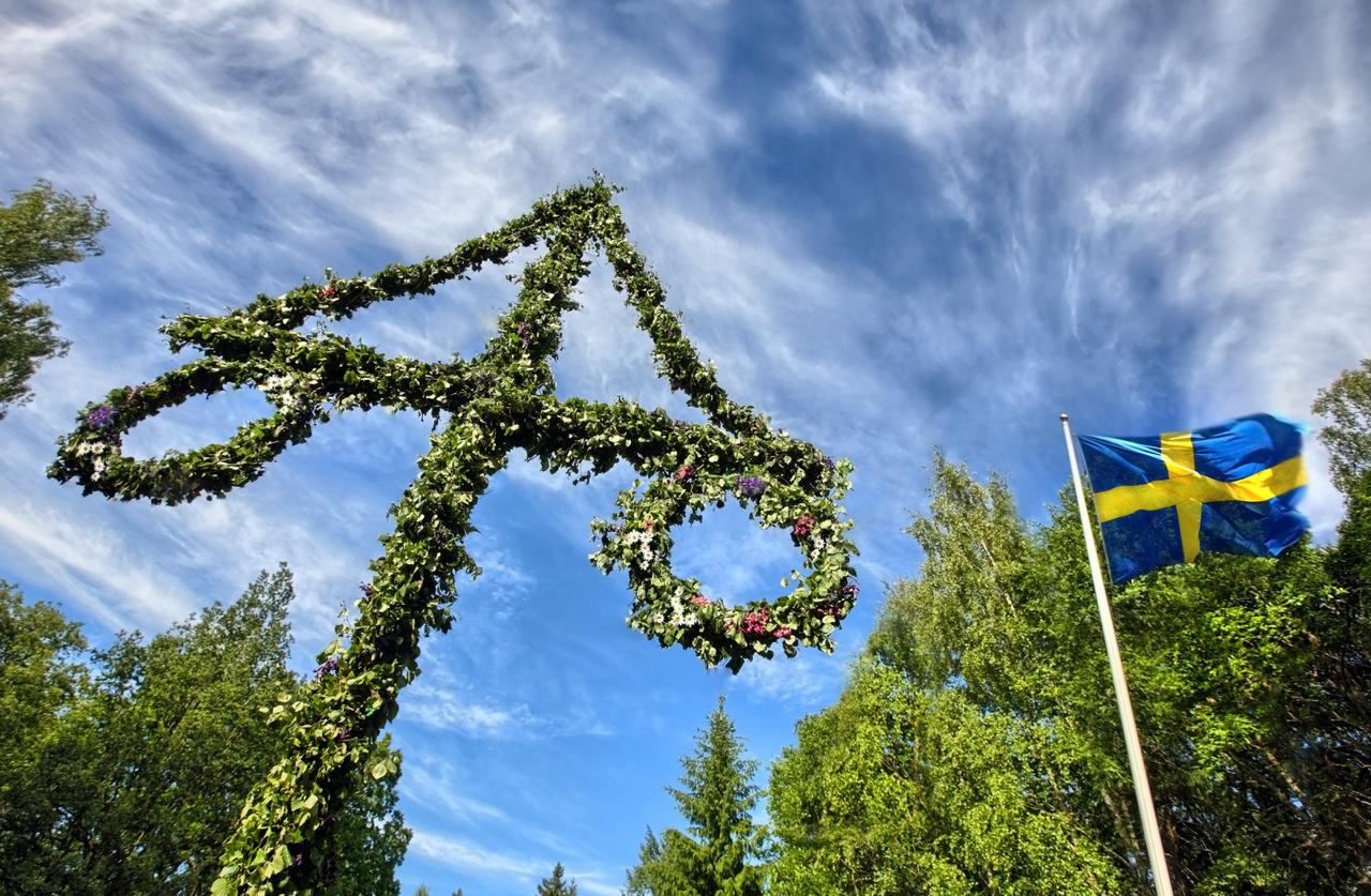 Midsommarstång och svensk flagga tornar upp sig mot en blå himmel.
